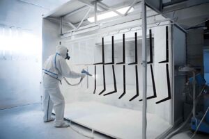 A worker performing the powder coating process by spray-coating metal angle parts inside a closed powder coating booth.